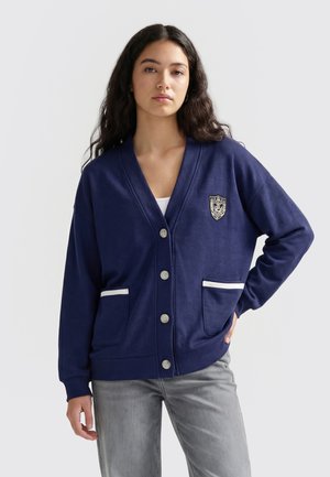 Woman wearing a navy blue buttoned cardigan with white-trimmed pockets and an embroidered crest, paired with light gray jeans.