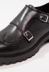 Chaussure à double boucle moniale en cuir noir avec boucles argentées, détails brogue et semelle épaisse à crampons.