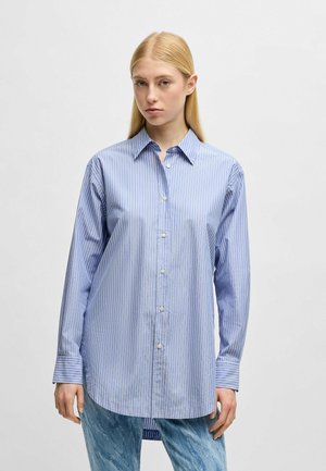 Camicia a righe azzurra chiara con righe verticali bianche. Presenta un colletto e maniche lunghe con polsini a bottoni. Vestibilità aderente.