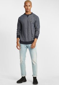 Tengeri kék texturált pulóver, raglán ujjakkal, fekete gallérral és egy kis logóval; világoskék, szűkített farmerral és fekete lábbelivel párosítva.