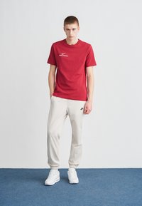 T-shirt vermelha de algodão com estampa branca "Nike Official", combinada com calças de fato de treino bege claras com logótipo preto e riscas laterais brancas. Ténis brancos.