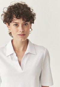 Bílá košile s krátkým rukávem; vyrobená z měkké látky s jemným leskem. Modelka má přirozený make-up a náušnice ve tvaru kruhu.