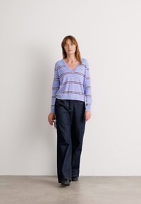 Pull léger violet en V avec des rayures horizontales, associé à un pantalon large en denim foncé et des chaussures noires, créant un look décontracté.