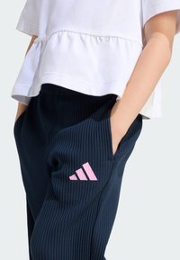 Pantalones azul marino acanalados con logo rosa de Adidas, combinados con una blusa blanca peplum. El conjunto muestra bolsillos y una silueta relajada.