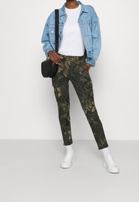 Pantalon cargo camoufflé avec motif floral vert et accents ; assorti à une veste en jean bleu clair, un t-shirt blanc et des baskets blanches.