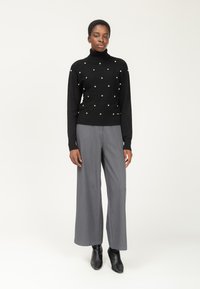 Schwarzer Pullover mit hohem Rollkragen, verziert mit silbernen Nieten in einem Diamantmuster. Kombiniert mit grauen weit geschnittenen Hosen und schwarzen Knöchelstiefeln.