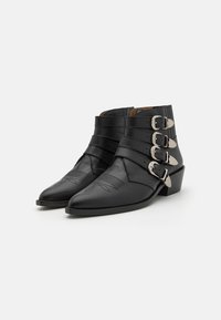 Botine din piele neagră până la gleznă, cu vârfuri ascuțite, prezentând multiple curele și catarame de argint, detalii embosate pe vârf și o talpă joasă.