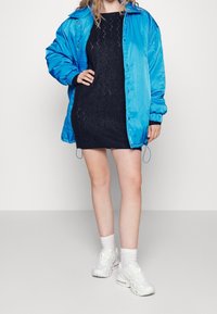 Veste oversize satinée bleue sur une robe tricotée noire à motif géométrique, associée à des baskets blanches et des chaussettes courtes.