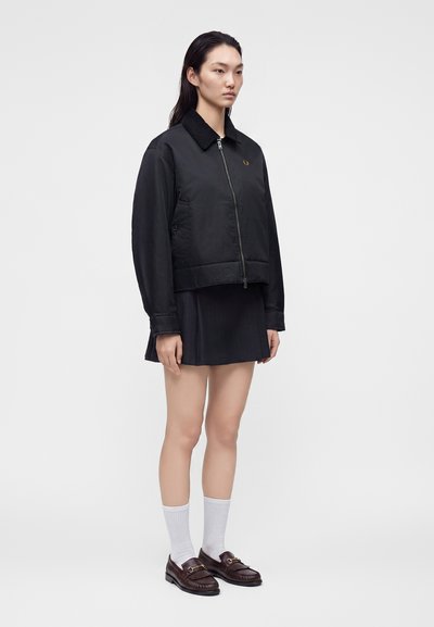 Jeune femme aux longs cheveux foncés porte une veste zippée noire, une jupe noire, des chaussettes blanches et des mocassins marron foncé, debout devant un fond uni.