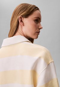 Jeune femme aux cheveux châtain clair et raides, portant une chemise à col rayée jaune et blanche, regardant vers la droite sur un fond gris.