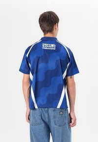 Short-sleeve, blue sports shirt with wavy pattern, white accents, and "OBEY SONIC VIBRATIONS" graphic on the back. Lightweight fabric.