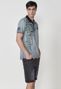 Siv polo majica z zelenim grafičnim tiskom in črnim ovratnikom, v kombinaciji z temnimi denim kratkimi hlačami s zavihanimi robovi in subtilnimi detajli.