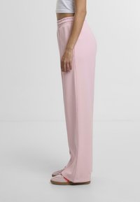 Lyserøde, brede sweatpants med elastisk talje, lavet af blødt materiale. Kombineret med lyserøde sneakers med orange detaljer.