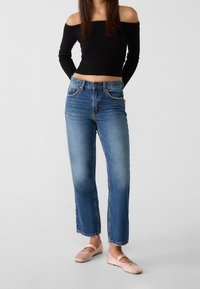 Mujer vestida con una blusa negra de manga larga y hombros al aire, jeans rectos azul de cintura alta y bailarinas rosas claras, de pie con las manos detrás de la espalda.