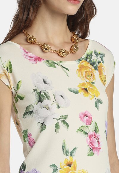 Frau trägt ein weißes Blumenkleid mit gelben, rosa und violetten Blumen sowie eine grobe goldene Kettenhalskette mit ineinander verflochtenen Knoten.