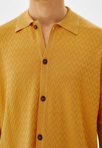 Żółty sweter w serek z krótkim rękawem w zygzakowaty wzór, z prążkowanym kołnierzem i ciemnobrązowymi guzikami na przodzie.
