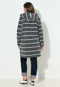 Cappotto in pile a righe grigie con cappuccio, caratterizzato da righe bianche, una vestibilità ampia e una spacco laterale. Indossato sopra jeans scuri e scarpe bianche.