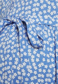 Tecido azul com um acabamento texturizado, apresentando uma estampa floral branca. Um detalhe de cinto atado é visível, criando um visual estruturado.