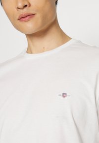 Κοντινό πλάνο ενός ατόμου που φοράει μια απλή λευκή μπλούζα με στρογγυλή λαιμόκοψη και ένα μικρό κεντητό λογότυπο σε κόκκινο, λευκό και μπλε στο αριστερό στήθος.