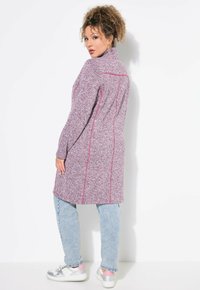 Fitted lang cardigan i en meleret lilla strik, med pink syninger, høj krave og sidelommer, parret med lyseblå jeans.