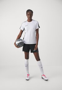 Hvit Nike T-skjorte med svarte detaljer, svarte Nike shorts, hvite sokker og fotballsko med røde detaljer; holder en metallisk fotball.