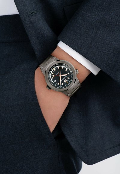 Reloj con una caja redonda de acero inoxidable plateado, esfera negra con marcadores blancos y naranjas, pulsera de metal, mostrado sobre la manga de un traje oscuro.