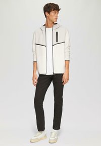 Lichtgrijze capuchonjack met zwarte accenten en een ritszak, gecombineerd met een witte t-shirt en zwarte jeans; model draagt lichtbeige sneakers.
