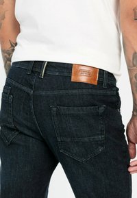 Donkerblauwe denim jeans met een soepele textuur, voorzien van achterzakken en een bruine leren label met de opdruk "Camel Active."