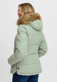 Chaqueta de plumas verde claro con capucha forrada de piel, diseño acolchado y detalles de botones a presión en los puños. Textura suave y corte informal.