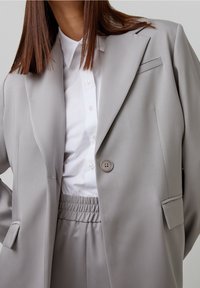 Helle graue Anzugjacke mit Spitzenrevers, Ein-Knopf-Verschluss und eingeschnittenen Taschen; getragen über einem weißen Hemd mit Button-Down-Kragen.