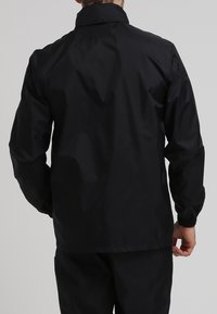 Mies, jolla on päällä yksinkertainen musta tuulitakki ja mustat housut, näkyy selkäpuolelta neutraalia taustaa vasten.