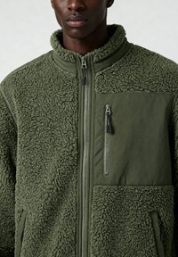 Veste polaire verte avec une surface texturée et duveteuse ainsi qu'un panneau en tissu lisse. Comprend une poche zippée et un col montant.