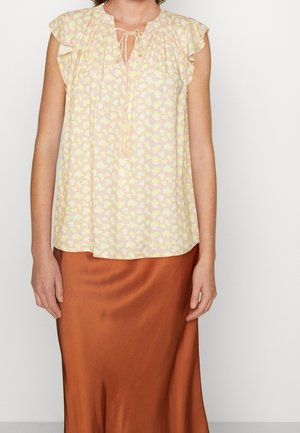 Blusa floral amarilla clara con mangas cortas acampanadas, escote fruncido y detalle de lazo, combinada con una falda de satén marrón.