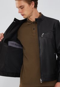 Zwarte leren jas met een rits aan de voorkant, ribbeldetails op de schouders en een borstzak. Draagt over een bruine polo.
