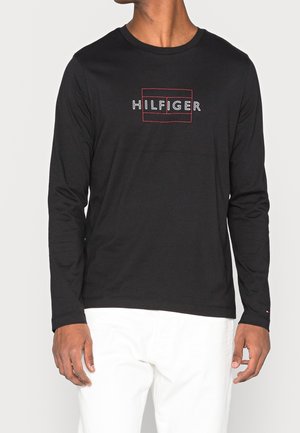 Mężczyzna ubrany w czarną koszulę z długim rękawem z napisem "HILFIGER" obrysowanym na czerwono na piersi oraz w białe spodnie, na jednolitym tle.