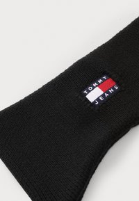 Bonnet en tricot noir avec un patch rectangulaire de logo Tommy Jeans en broderie bleu marine, blanc et rouge sur le côté. Texture côtelée simple.