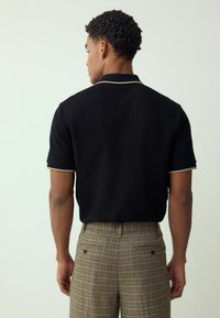 Schwarzes Polohemd mit kurzen Ärmeln, strukturierter Stoff, kontrastierende Kragenkanten. Kombiniert mit karierter Hose mit Gesäßtaschen.