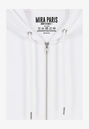 Balts kokvilnas rāvējslēdzēja džemperis ar sudraba rāvējslēdzēju, regulējamām baltiem auklām un etiķeti ar kopšanas instrukcijām.