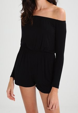 Femme portant une combinaison-short noire à épaules dénudées et manches longues avec une taille élastique, debout devant un fond clair uni.