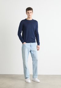 Sötétkék, hosszúujjú pulóver kötött anyagból, világoskék, egyenes szárú farmerral és fehér sportcipővel egy semleges háttér előtt.