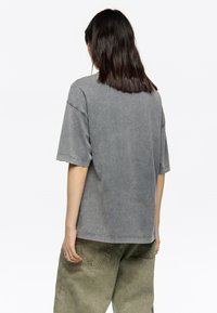 Persona con cabello oscuro hasta los hombros, vistiendo una camiseta gris deslavada de gran tamaño y pantalones sueltos color verde oliva, de espaldas sobre un fondo blanco.