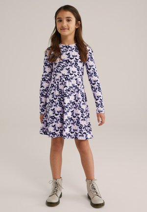 Junges Mädchen steht mit einem langärmeligen lavendelfarbenen Kleid mit maritimen Blumenmuster und hellbeigen Schnürstiefeletten vor einem schlichten Hintergrund.