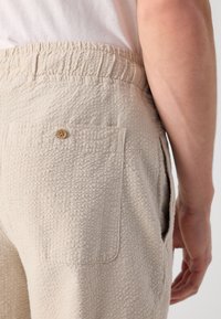 Gros plan sur la taille et le bras d'une personne portant un pantalon beige texturé avec une ceinture élastique et une poche arrière sécurisée par un petit bouton.