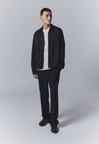 Chaqueta negra con cremallera sobre una camiseta blanca, combinada con pantalones negros y zapatos negros. Diseño simple con un corte relajado.