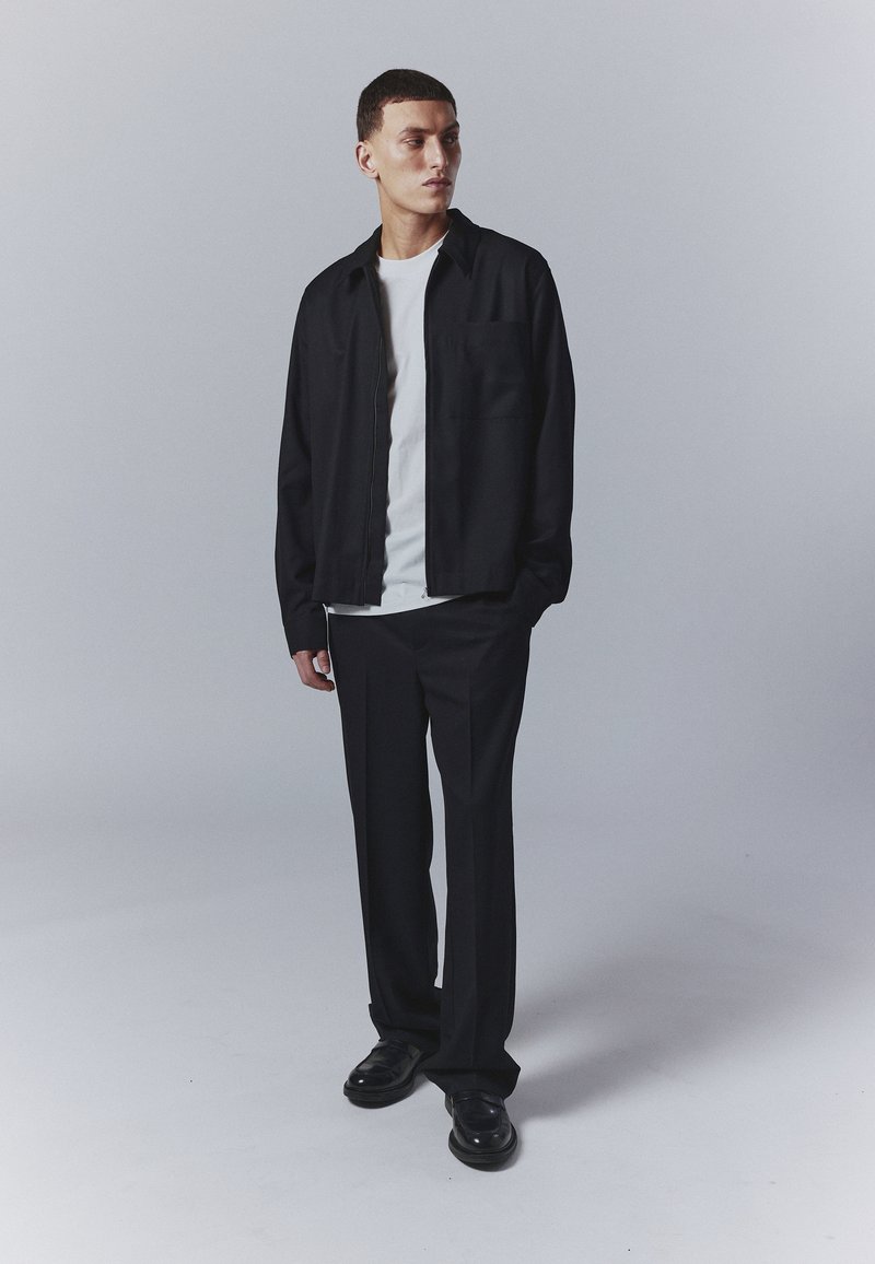 Sort zip-jakke over en hvid t-shirt, parret med sorte bukser og sorte sko. Enkel design med en afslappet pasform.