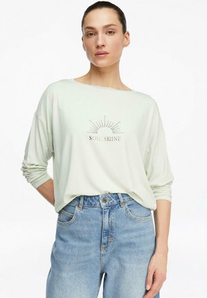 Kobieta w jasnozielonej koszuli z długim rękawem z napisem "SOUL SHINE" i grafiką słońca, w połączeniu z wysokim stanem niebieskich dżinsów, stojąca na białym tle.