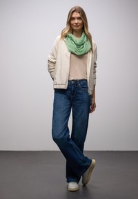 Haut beige, veste zippée crème clair, écharpe verte à motifs, jean flare bleu, baskets blanches, posture debout sur un sol gris, fond neutre.