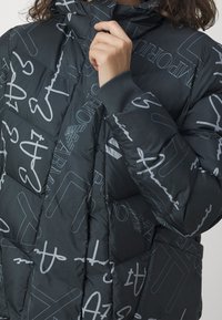 Person som bär en svart vadderad jacka med vita skript- och logotyp mönster, som justerar kragen nära hakan.