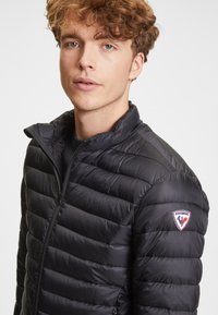 Mladý muž s kudrnatými vlasy, oblečený v černé prošívané bundě Rossignol se zapínacím límcem, který se dívá mírně mimo kameru.