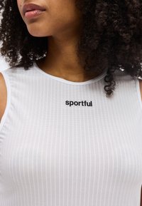 Εγγύς πλάνο ενός ατόμου με σγουρά μαλλιά που φοράει λευκή αμάνικη μπλούζα με λεπτές κάθετες ρίγες και τη λέξη «sportful» σε μαύρο κείμενο.